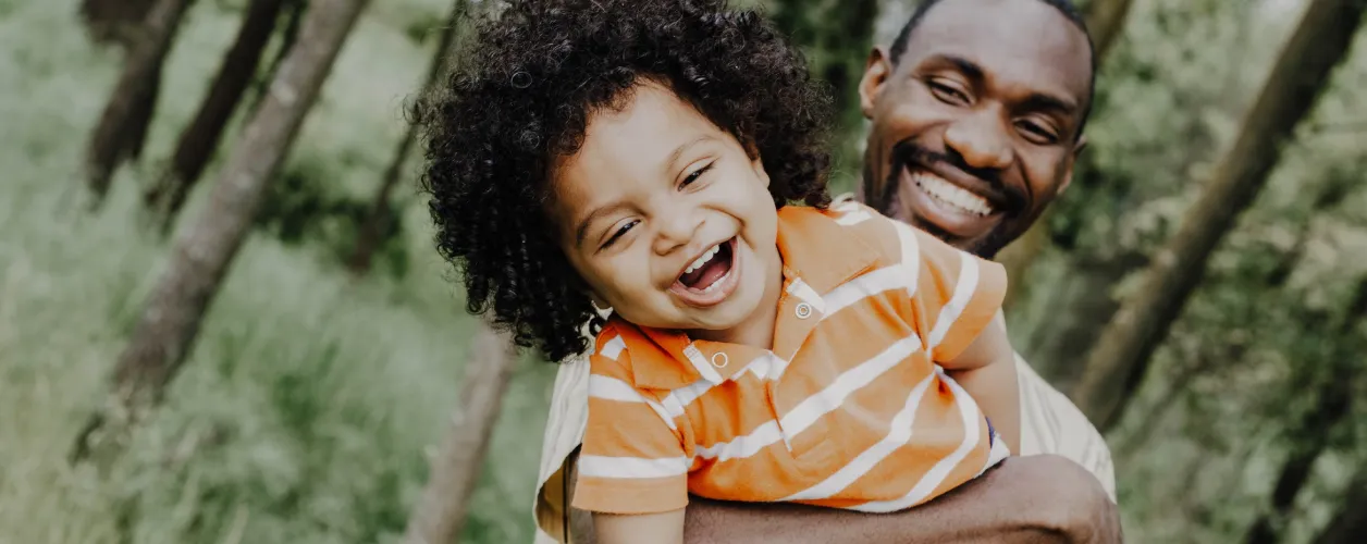 A caregiver laughing with a joyful young child on his back, outdoors among trees